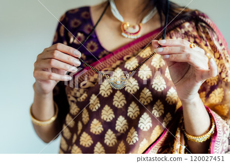 Dressed in beautiful saree, Indian woman delicately putting traditional jewellery necklace getting ready for ethnic festive celebration. Indian culture, ritual holiday in modern life. Selective focus. 120027451