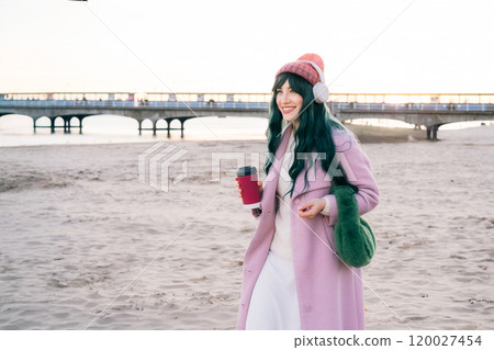 Stylish young smiling hipster woman with color hair wearing wireless headphones, pink coat, hat, walking on beach with reusable coffee cup. Time To Relax. seasonal fashion style trend. Barbiecore. Stylish young smiling hipster woman with color hair wearing wireless headphones, pink coat, hat, walking on beach with reusable coffee cup. Time To Relax. seasonal fashion style trend. Barbiecore. 120027454
