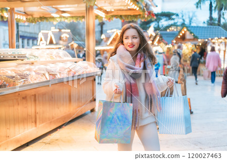 Happy woman enjoys shopping at festive Christmas market while carrying shopping paper bags, walking in street Xmas fair of European city. Selecting gifts for New Year, winter holiday sales, discounts. Happy woman enjoys shopping at festive Christmas market while carrying shopping paper bags, walking in street Xmas fair of European city. Selecting gifts for New Year, winter holiday sales, discounts. 120027463