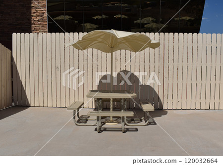 Empty benches and a table under parasol, beige sun umbrella against the background of a beige fence Empty benches and a table under parasol, beige sun umbrella against the background of a beige fence 120032664