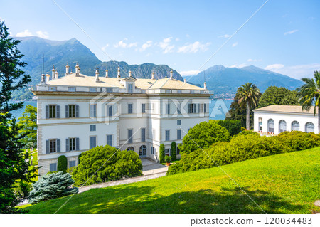 The elegant Villa Melzi overlooks Lake Como, surrounded by lush gardens and majestic mountains, inviting visitors to appreciate its beauty in the warm summer sunshine. 120034483