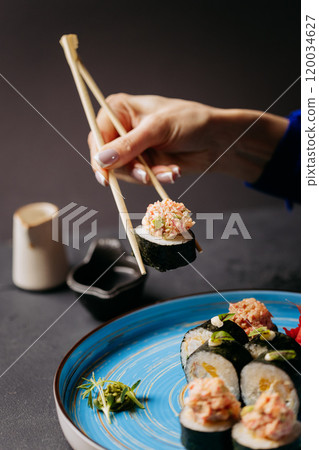 Hand using chopsticks to hold sushi roll with crab topping, served on a blue plate with soy sauce Hand using chopsticks to hold sushi roll with crab topping, served on a blue plate with soy sauce 120034627