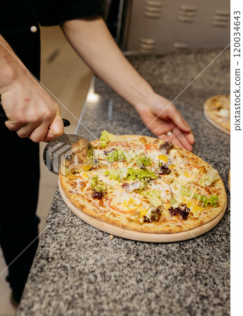 Chef slicing fresh vegetable pizza with pizza cutter on granite countertop for a perfect serving 120034643