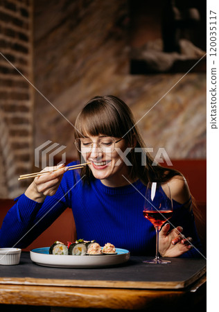 Smiling woman enjoying sushi with chopsticks, paired with a glass of rose wine in a cozy restaurant 120035117