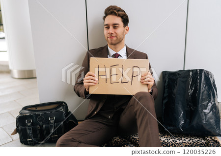 Portrait of poor handsome millennial businessman in suit sitting on city street holding sign HELP, illustrating hardships of poverty and unemployment. Concept of support in financial crises. 120035226