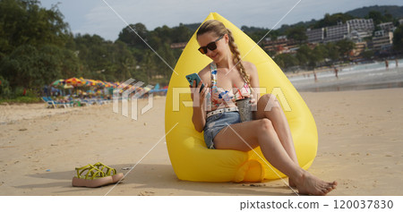 A young woman on the beach lounging on a yellow bean bag  120037830