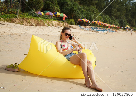A young woman on the beach lounging on a yellow bean bag  120037869