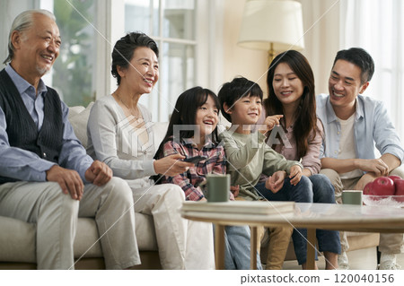 three generation asian family watching tv together at home 120040156