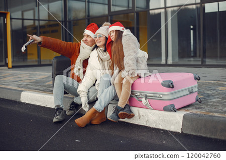 Family of three going on vacation. Man and girl sitting on a luggage. Mother, father and daughter wearing warm clothes and santa claus hats. Family of three going on vacation. Man and girl sitting on a luggage. Mother, father and daughter wearing warm clothes and santa claus hats. 120042760