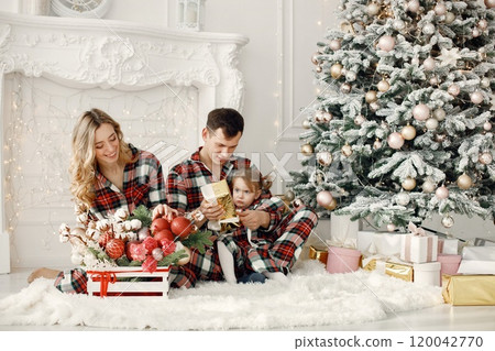Blonde mother, brunette father and little daughter sitting on a floor near Christmas tree. Woman and daughter wearing plaid pajamas. Family opening a Christmas gifts together. 120042770