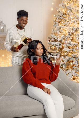 Young black couple with gifts in Christmas eve. Woman sitting on a coach at home. Man standing behind and presented a gift for his girlfriend. Young black couple with gifts in Christmas eve. Woman sitting on a coach at home. Man standing behind and presented a gift for his girlfriend. 120042802