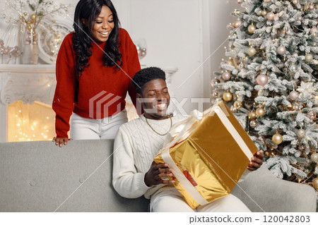 Young black couple with gifts in Christmas eve. Man sitting on a coach at home. Brunette woman standing behind and presented a gift for her boyfriend. 120042803