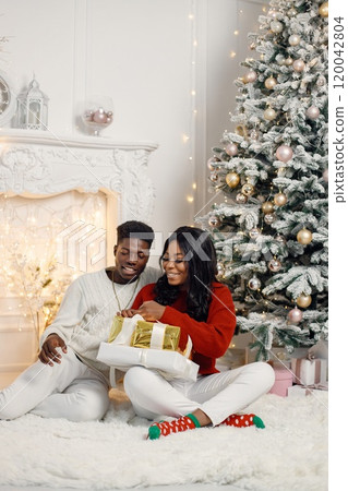 Happy black couple holding gifts and sitting near Christmas tree. Man amd woman celebrating winter holidays at home. Brunette woman wearing a red sweater. Happy black couple holding gifts and sitting near Christmas tree. Man amd woman celebrating winter holidays at home. Brunette woman wearing a red sweater. 120042804