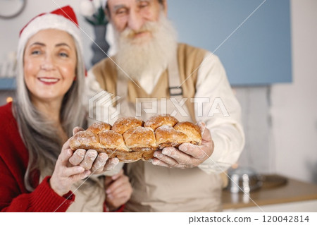 Senior man and his wife baking bread together. Old bearded man standing together near his wife with grey hair. Man wearing white sweater and woman red one. Senior man and his wife baking bread together. Old bearded man standing together near his wife with grey hair. Man wearing white sweater and woman red one. 120042814