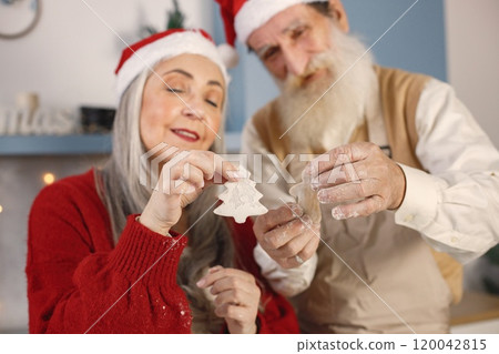 Senior man and his wife baking cookies together. Old bearded man standing together near his wife with grey hair. Man wearing white sweater and woman red one. Senior man and his wife baking cookies together. Old bearded man standing together near his wife with grey hair. Man wearing white sweater and woman red one. 120042815