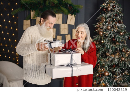 Senior man celebrating christmas with his wife near beautiful christmas tree. Old bearded man standing near his wife with grey hair and holding a gifts. Man wearing white sweater and woman red one. 120042819