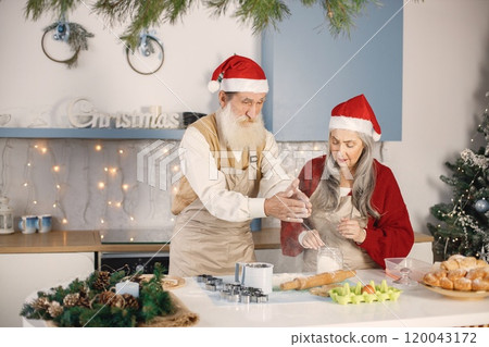 Senior man and his wife cooking together. Old bearded man standing together near his wife with grey hair. Man wearing white sweater and woman red one. Senior man and his wife cooking together. Old bearded man standing together near his wife with grey hair. Man wearing white sweater and woman red one. 120043172