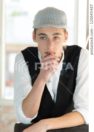 A young man in a cap, a vest, a white shirt, smokes by the window and talks. Bad habits among young people. Young manager. A young man in a cap, a vest, a white shirt, smokes by the window and talks. Bad habits among young people. Young manager. 120045487