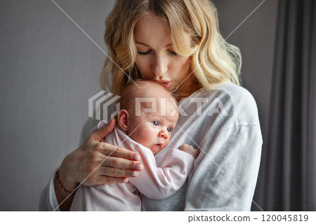 Beautiful young blonde mother kissing baby, dressed in light clothing indoor. Mom and newborn on grey background, Tender loving connection between mom and child, care and affection in peaceful 120045819