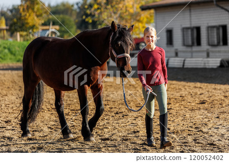 Young little teenager kid girl leading chestnut horse through stable ranch yard. Horse riding school exercise. Cute little beginner blond girl kid near beautiful brown horse. Equine hobby or work Young little teenager kid girl leading chestnut horse through stable ranch yard. Horse riding school exercise. Cute little beginner blond girl kid near beautiful brown horse. Equine hobby or work 120052402
