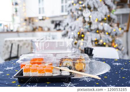 Delivery of sushi and rolls in food containers on the table in the festive interior of the kitchen of the house with a white Christmas tree for Christmas and New year. Food delivery for the holidays 120053285