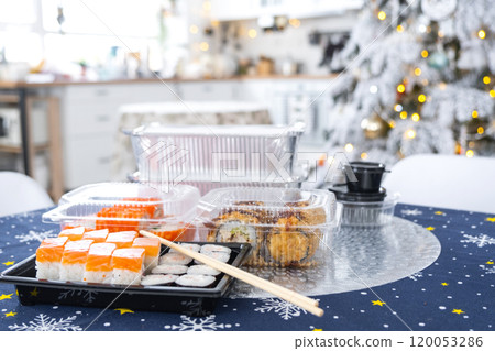 Delivery of sushi and rolls in food containers on the table in the festive interior of the kitchen of the house with a white Christmas tree for Christmas and New year. Food delivery for the holidays 120053286