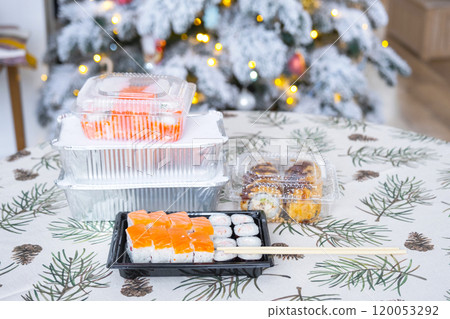 Delivery of sushi and rolls in food containers on the table in the festive interior of the kitchen of the house with a white Christmas tree for Christmas and New year. Food delivery for the holidays 120053292