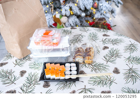 Delivery of sushi and rolls in food containers on the table in the festive interior of the kitchen of the house with a white Christmas tree for Christmas and New year. Food delivery for the holidays 120053295