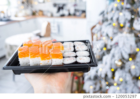 Delivery of sushi and rolls set is hold in hand in the festive interior of house with a white Christmas tree for Christmas and New year. Food delivery for the holidays 120053297