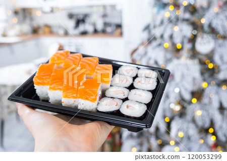 Delivery of sushi and rolls set is hold in hand in the festive interior of house with a white Christmas tree for Christmas and New year. Food delivery for the holidays 120053299