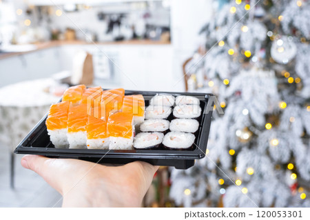 Delivery of sushi and rolls set is hold in hand in the festive interior of house with a white Christmas tree for Christmas and New year. Food delivery for the holidays Delivery of sushi and rolls set is hold in hand in the festive interior of house with a white Christmas tree for Christmas and New year. Food delivery for the holidays 120053301