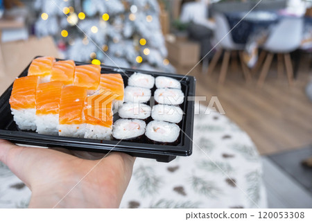 Delivery of sushi and rolls set is hold in hand in the festive interior of house with a white Christmas tree for Christmas and New year. Food delivery for the holidays 120053308