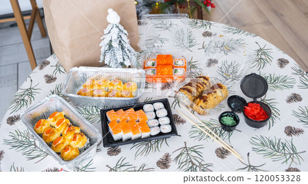 Delivery of sushi and rolls set top view on the table with a Christmas decor with a Christmas tree and a tablecloth with snowflakes, home cooking 120053328
