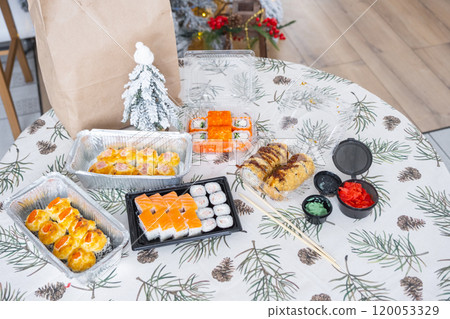 Delivery of sushi and rolls in food containers on the table in the festive interior of the kitchen of the house with a white Christmas tree for Christmas and New year. Food delivery for the holidays 120053329