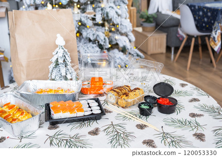 Delivery of sushi and rolls in food containers on the table in the festive interior of the kitchen of the house with a white Christmas tree for Christmas and New year. Food delivery for the holidays Delivery of sushi and rolls in food containers on the table in the festive interior of the kitchen of the house with a white Christmas tree for Christmas and New year. Food delivery for the holidays 120053330