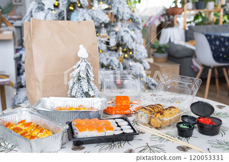 Delivery of sushi and rolls in food containers on the table in the festive interior of the kitchen of the house with a white Christmas tree for Christmas and New year. Food delivery for the holidays Delivery of sushi and rolls in food containers on the table in the festive interior of the kitchen of the house with a white Christmas tree for Christmas and New year. Food delivery for the holidays 120053331