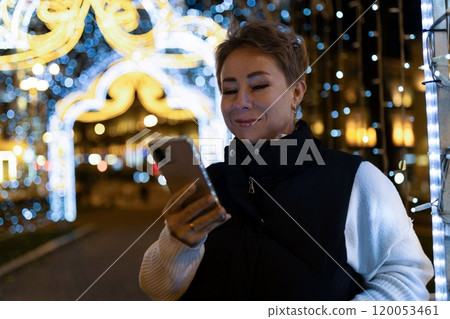 A woman walks through a city at night, looking at her phone, with bright lights illuminating the A woman walks through a city at night, looking at her phone, with bright lights illuminating the 120053461