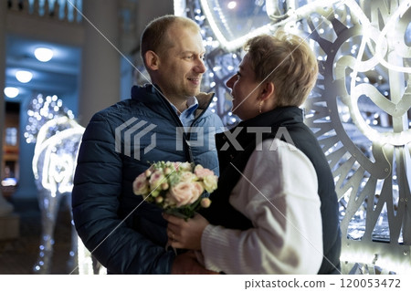 A couple walks through a city at night, the man holding a bouquet of flowers 120053472