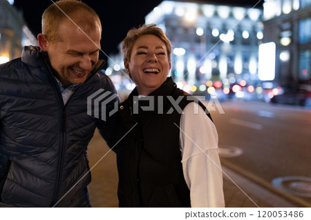 A couple walks through the city at night, laughing as they enjoy their evening A couple walks through the city at night, laughing as they enjoy their evening 120053486