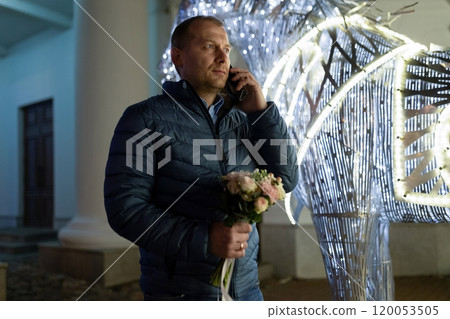 A man walks through the city at night, holding a bouquet of flowers and talking on his phone A man walks through the city at night, holding a bouquet of flowers and talking on his phone 120053505