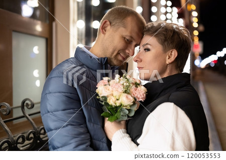 A couple walks through the city at night, holding a bouquet of pink flowers A couple walks through the city at night, holding a bouquet of pink flowers 120053553