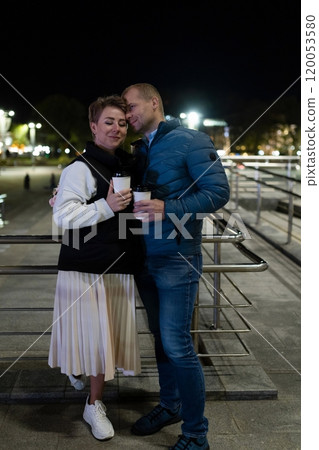 A couple strolls through the city at night, holding warm drinks as they enjoy each other's company 120053580