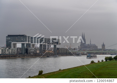 A modern cityscape featuring three unique crane-shaped buildings along the river, with a bridge in front carrying a tram. View of Cologne Cathedral with a passenger boat navigating the Rhine River. A modern cityscape featuring three unique crane-shaped buildings along the river, with a bridge in front carrying a tram. View of Cologne Cathedral with a passenger boat navigating the Rhine River. 120054867