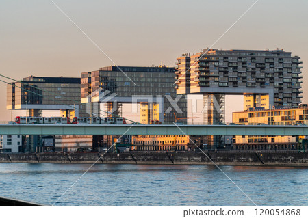 A modern cityscape featuring three unique crane-shaped buildings along the river, with a bridge in front carrying a train. The buildings reflect the warm light of the setting sun. 120054868