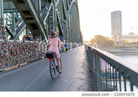 A woman on a bicycle rides on a bridge over the Rhine in the city of Cologne. A beautiful metal bridge at dawn. Many locks are attached to the bridge. The woman in a red dress and a red helmet. 120054870