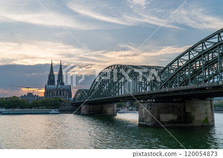 View of Cologne Cathedral and Hohenzollern Bridge with a cargo ship and passenger boat navigating the Rhine River under a clear blue sky, showcasing Cologne's iconic cityscape and vibrant river life. 120054873