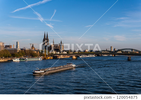 View of Cologne Cathedral and Hohenzollern Bridge with a cargo ship and passenger boat navigating the Rhine River under a clear blue sky, showcasing Cologne's iconic cityscape and vibrant river life. 120054875