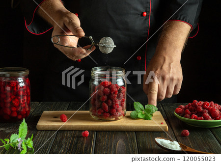 A chef skillfully uses a sieve to sprinkle sugar over a jar filled with fresh raspberries. Mint leaves and extra berries are arranged on the table, creating a warm, inviting atmosphere A chef skillfully uses a sieve to sprinkle sugar over a jar filled with fresh raspberries. Mint leaves and extra berries are arranged on the table, creating a warm, inviting atmosphere 120055023