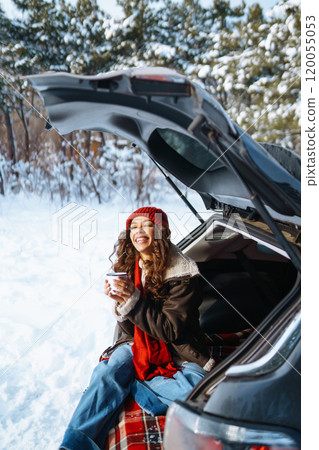 Young woman holds a thermos and drinks tea sitting in car trunk in sunny winter forest. Rest concept Young woman holds a thermos and drinks tea sitting in car trunk in sunny winter forest. Rest concept 120055053