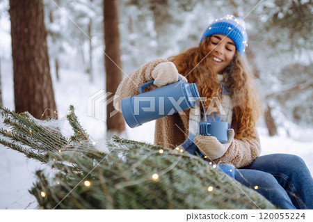 Young woman drinks a hot drink from a thermos while sitting in a snowy forest with a Christmas tree. 120055224